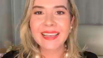 Dani Siriani dá seu depoimento e maravilhosa experiência sobre hipnose para burnout. Ela é loira, veste uma camisa preta e lindo colar de pérolas