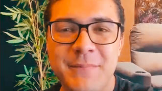 Lucas veste uma camisa preta, usa óculos e tem cabelos curtos encaracolados. Ele faz seu depoimento sobre sua experiência co a hipnoterapia para tratar uma raiva constante