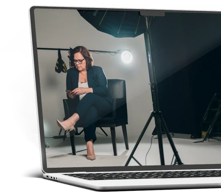 A foto mostra a tela de um notebook, com a foto da Denise Braga em um estúdio de gravação. Ela vez conjunto de blazer azul marinho e blusa branca.
