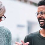 Dois amigos de trabalho conversam animadamente sobre um projeto. Ela tem cabelos curtos, está de óculos pretos e tailer azul claro. Ele está de camiseta preta, tem barba e cabelos pretos. Expressam a comunicação do amor entre colegas de trabalho