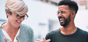 Dois amigos de trabalho conversam animadamente sobre um projeto. Ela tem cabelos curtos, está de óculos pretos e tailer azul claro. Ele está de camiseta preta, tem barba e cabelos pretos. Expressam a comunicação do amor entre colegas de trabalho