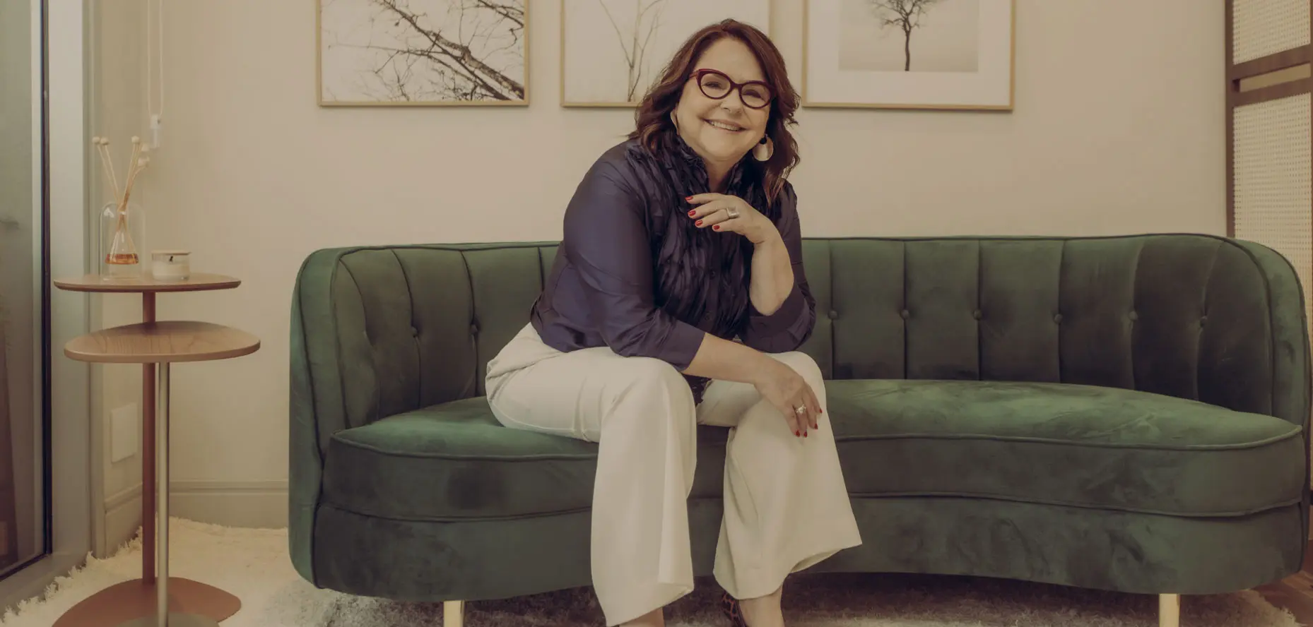 Denise Braga está sentada em um sofá verde, super moderno. Ela veste camisa de babados azul de seda e calça bege. Ela está na sala da Casa 33