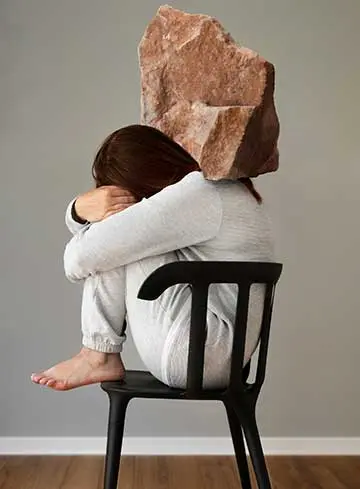 A imagem mostra uma moça sentada em uma cadeira, em posição fetal, com um peso simbôlico como uma pedra, representando a depressão