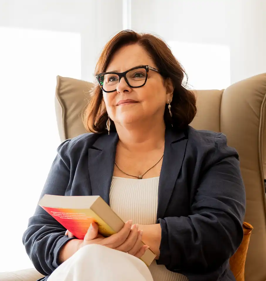 Denise Braga está sentada em uma poltrona, segurando um livro nas mãos. Ela veste blazer azul marinho, blusa e calças claras. Ela fala sobre os serviços de hipnoterapia