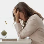 Mulher está sentada a mesa de um escritório, com as mãos no rosto. Ela demostra o adoecimento mental no trabalho