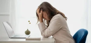 Mulher está sentada a mesa de um escritório, com as mãos no rosto. Ela demostra o adoecimento mental no trabalho