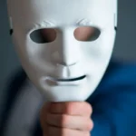 Homem de paletó e gravata, segura uma máscara na frente do seu rosto, à distância. Ele quer representar a Síndrome do Impostor