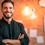 Homem de camisa escura, barba, está de braços cruzados, mostrando a força da sua transição para o empreendedorismo