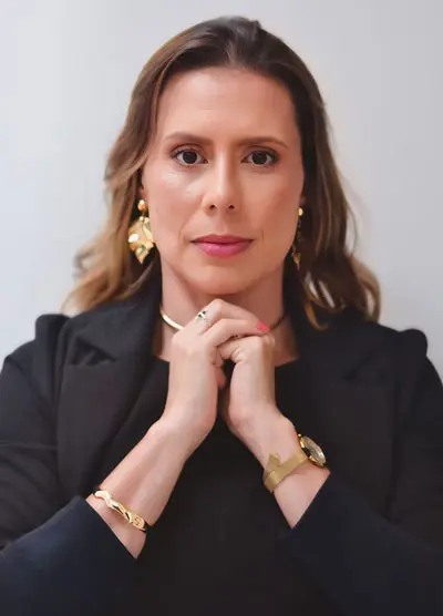Foto de Caroline Vasconcelos. Ela tem cabelos castanhos claros na altura dos ombros. Ela uma um vestido e um tailer azul marinho.