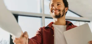 Um homem jovem de barba, camisa vinho e camiseta branca, está apertando a mão de uma pessoa que não aparece na foto. Essa foto representa o poder da consistência.