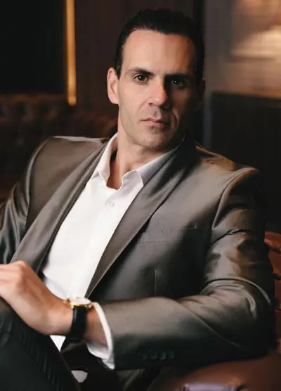Rodrigo Gonçalves está de terno em tom de marrom e camisa branca sem gravata. Ele está sentado em uma poltrona de couro marrom terra.