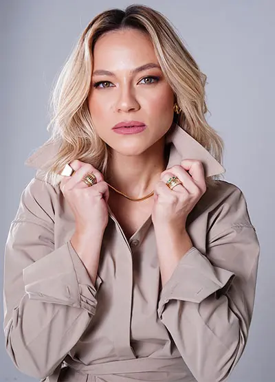 Foto da Samya Leão. Ela está de cabelo solto na altura dos ombros. Ela é loira e segura a gola da camisa com as duas mãos