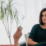 Uma mulher de cabelo chanel e castanho, está sentada em uma poltrona de encosto baixo. Ela faz uma sessão de hipnoterapia para reprogramação