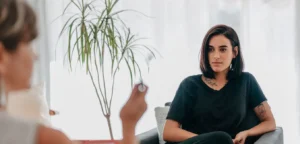 Uma mulher de cabelo chanel e castanho, está sentada em uma poltrona de encosto baixo. Ela faz uma sessão de hipnoterapia para reprogramação