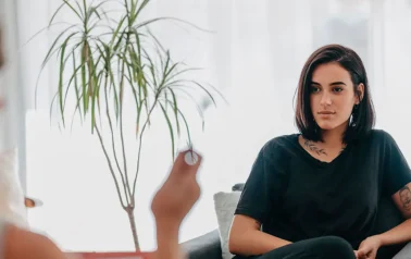 Uma mulher de cabelo chanel e castanho, está sentada em uma poltrona de encosto baixo. Ela faz uma sessão de hipnoterapia para reprogramação