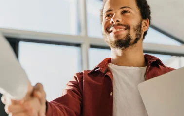 Um homem jovem de barba, camisa vinho e camiseta branca, está apertando a mão de uma pessoa que não aparece na foto. Essa foto representa o poder da consistência.